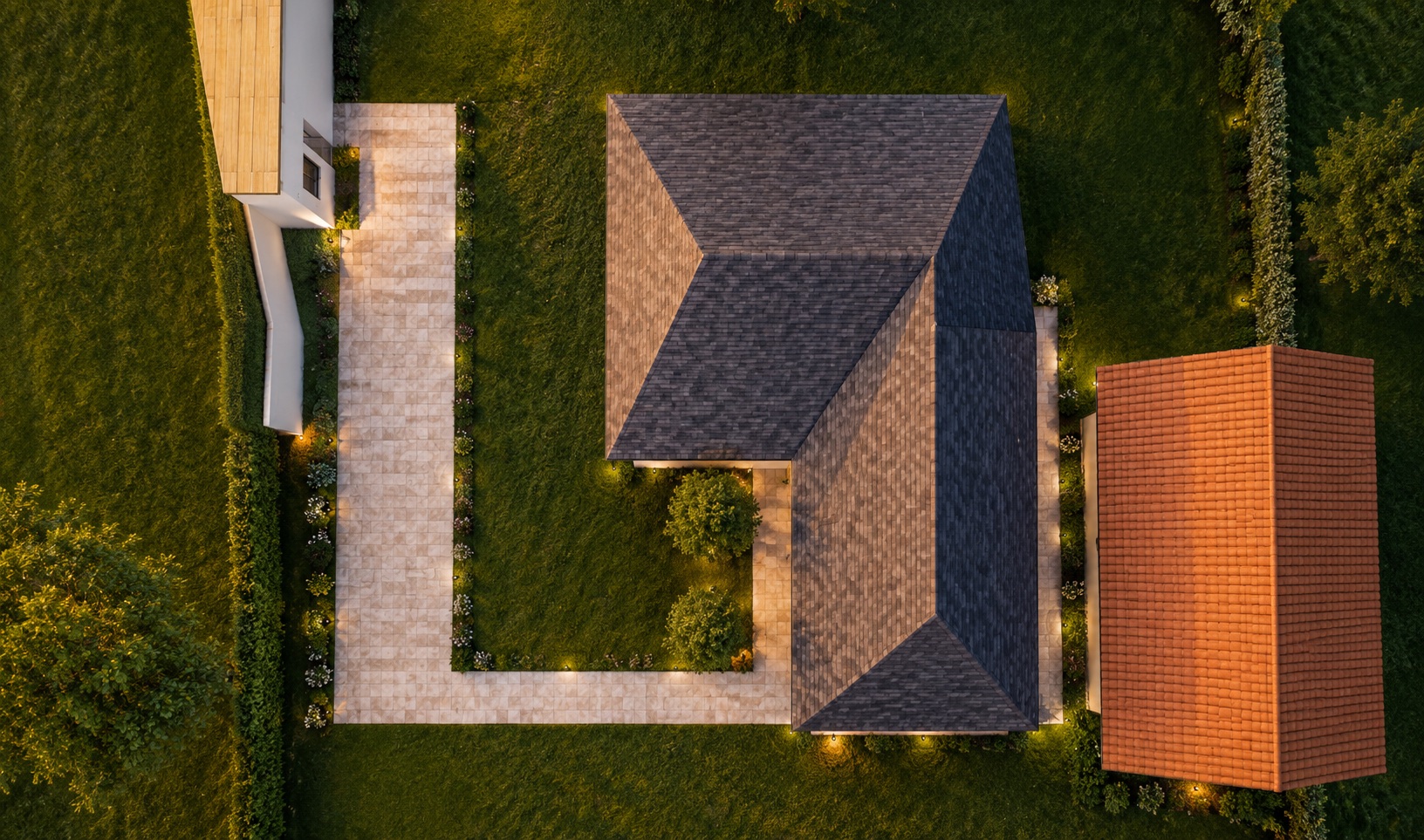Top-down view at dusk: two hipped roofs in dark shingle, a paved path wrapping the lawn, garden lights